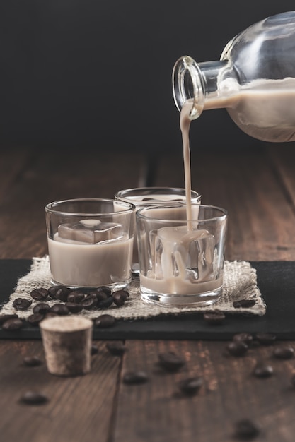 Pouring  Cream coffee liqueur in glasses with ice, surrounded by coffee beans on a dark wooden surface