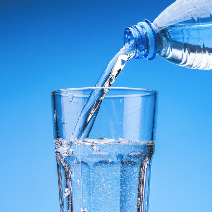 Photo pouring drinking water from plastic bottle into glass, against blue sky with clouds.