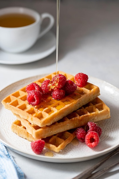 Pouring honey over sweet waffles with berries in plate served with cup of tea