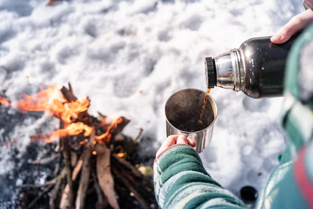 Pouring hot drink out of thermos at a campsite. Person getting warm near a campfire, point of view shot