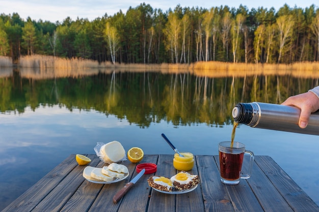 Pouring hot tea into a glass mug from a thermos in the morning next to the lake and forest in spring time, close up. Breakfast on a wooden table. Nature and travel concept