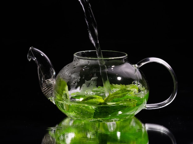 Photo pouring hot water into a glass teapot on a black background green mint tea herbal tea and healthy