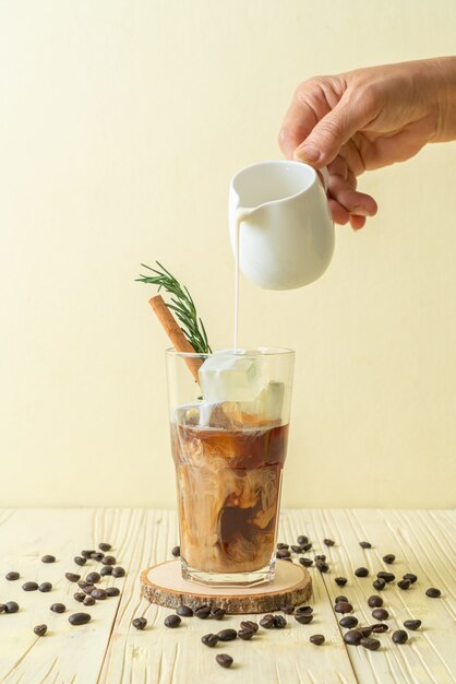 pouring milk in black coffee glass with ice cube, cinnamon and rosemary on wood surface