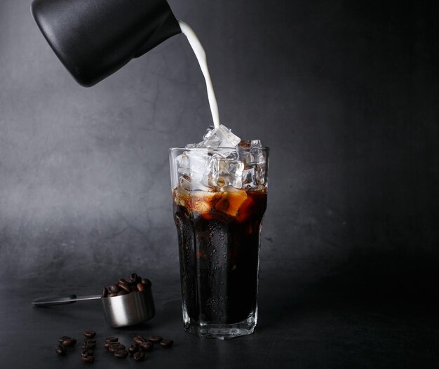 Pouring milk make iced latte coffee into a transparent glass\
and roasted coffee beans on a black background studio photox9