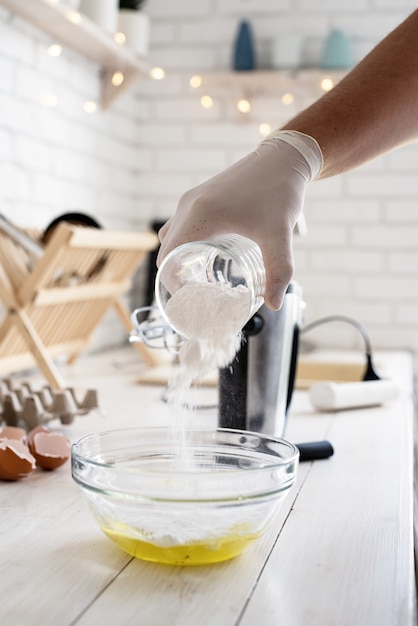 Pouring powder sugar into glass bow with eggs on white wooden table