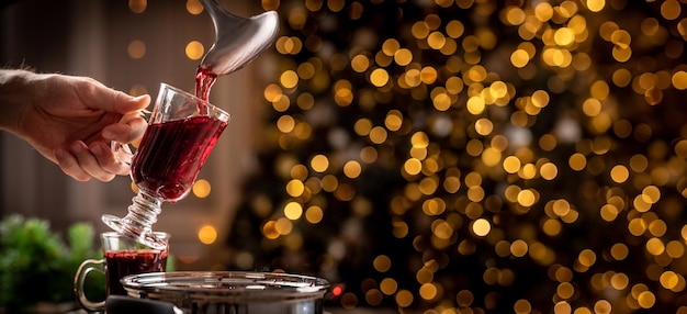 Pouring red mulled wine into transparent glass held by man on christmas illumination background