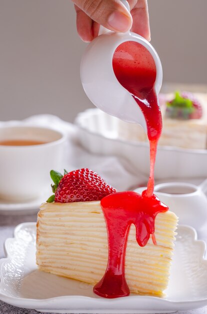 Pouring strawberry sauce onto delicious crepe cake on white plate.