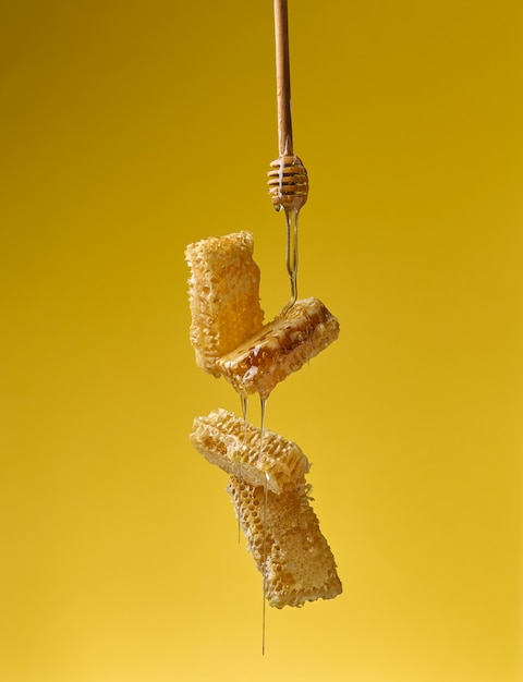 Pouring transparent sweet honey from a wooden stick on a wax\
honeycomb. yellow background. food levitates