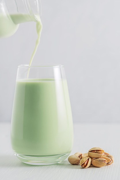 Pouring of vegan plant based pistachio milk from jar into glass served on white wooden table