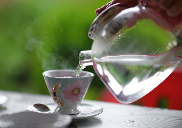 Pouring water from glass teapot into a cup on a blurred background of nature.