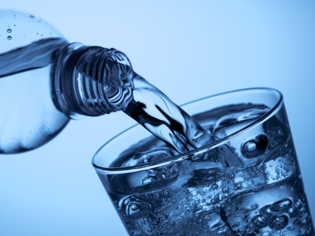 Pouring  water from a plastic bottle into a glass with ice on a light blue background