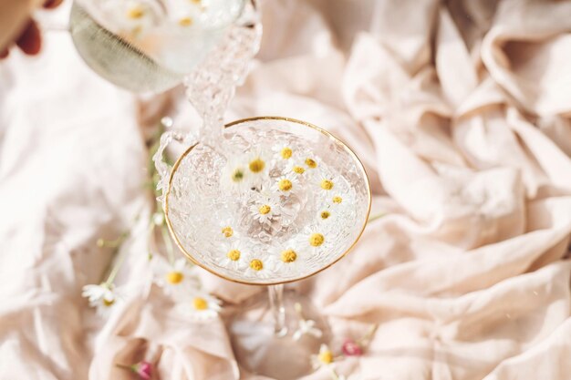 Pouring water with daisy flowers in modern wineglass on\
background of soft beige fabric with wildflowers tender floral\
aesthetic creative fresh summer drink bohemian mood