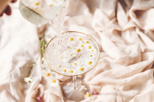 Pouring water with daisy flowers in modern wineglass on\
background of soft beige fabric with wildflowers tender floral\
aesthetic creative fresh summer drink bohemian mood