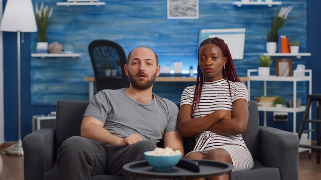 Photo pov of interracial couple being shocked watching television and drama movie at home. multi ethnic husband and wife looking at camera while sitting on couch in living room. mixed race people