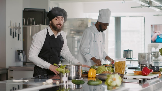 POV of male cook recording cooking show video on camera, explaining culinary food recipe. Young chef with uniform filming content for gastronomy TV program in professional kitchen. Tripod shot.