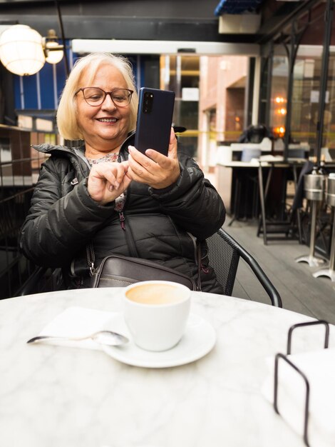 Foto pov oudere vrouw lacht en stuurt selfie's op haar mobiele telefoon met koffie die al klaar is.