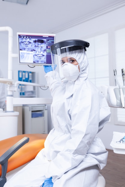 Photo pov of patient listening dentist talking about teeth hygine ponting at digital x-ray. stomatology specialist wearing protective suit against infection with coronavirus pointing at radiography.