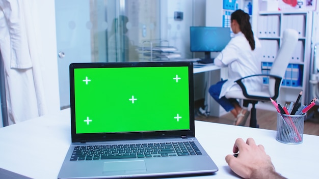 Pov shot of doctor using laptop with green screen in hospital cabinet. Medical worker wearing white coat in clinic. Medic using notebook with chroma key on display in medical clinic.