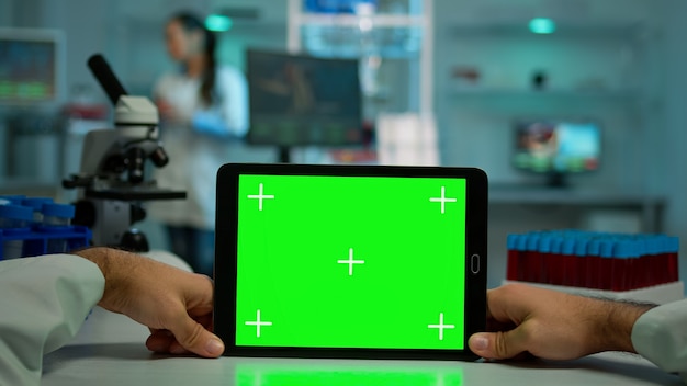 Pov shot of microbiologist holding tablet with green chroma key display sitting at desk working reading virus symptoms. In background lab researcher analysing vaccine developent examining samples