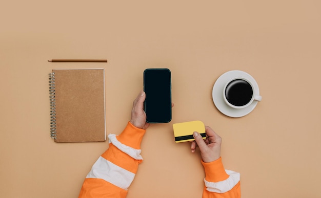 Photo pov view on female hands with smartphone next to notebook credit card and cup of coffee on brown background