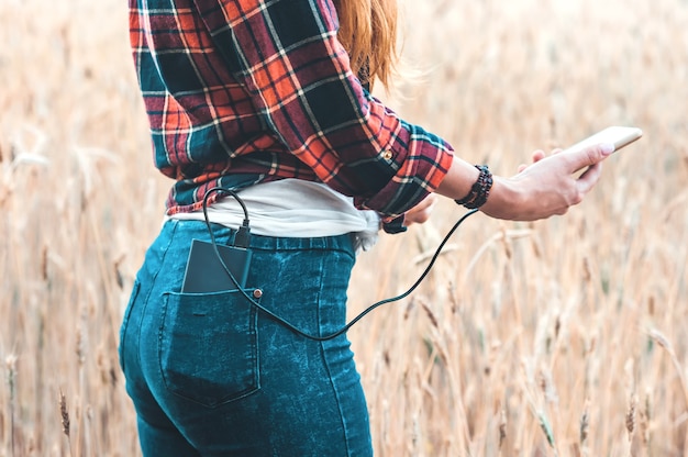 Power bank in the back pocket of a girl in a yellow field charges the phone, against the background of ears of wheat and sky, side view of a young beautiful woman.