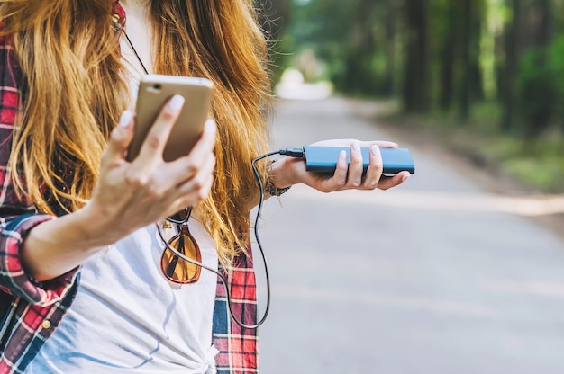 Power bank and smartphone in the hands of a girl with red hair in a shirt in a cage with a black backpack, on the background of a forest road.