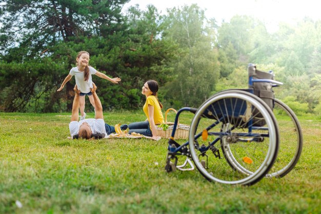 Power of imagination. Happy young man with disabilities lying on grass and lifting up his cheerful daughter as being an airplane while his wheelchair standing in the foreground