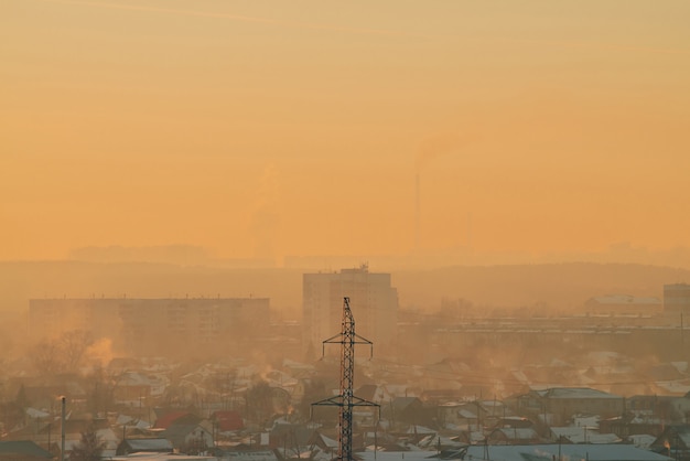 Power lines in city on dawn. Silhouettes of urban buildings among smog on sunrise. Cables of high voltage on warm orange yellow sky. Power industry at sunset. City power supply.