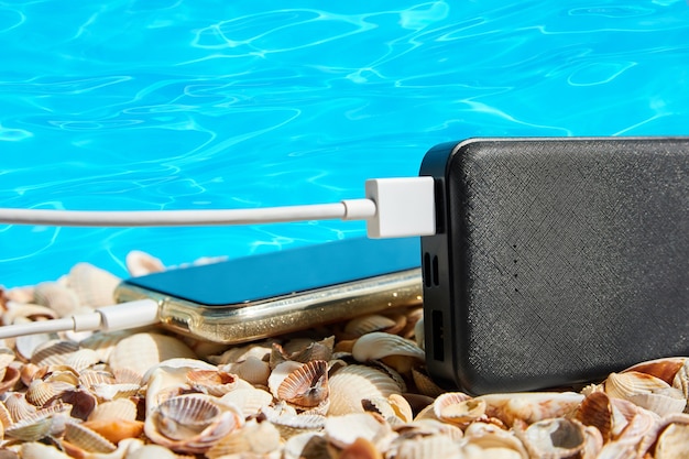 Powerbank charges a smartphone on seashells on blue water background