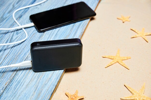 Powerbankは、夏の背景に木の板とヒトデの砂からスマートフォンを充電します