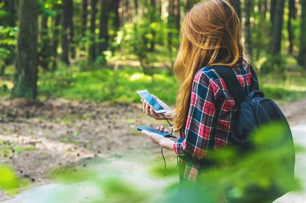 Powerbank en smartphone in de handen van een meisje met rood haar in een shirt in een kooi met een zwarte rugzak, op de achtergrond van een bosweg.