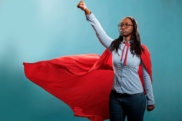 青い背景に飛んでいるようにポーズをとっている間、英雄の衣装を着ている強力で勇敢な若いスーパーヒーローの女性。カメラを見ながら野心的に見える誇り高きアフリカ系アメリカ人の正義の擁護者。