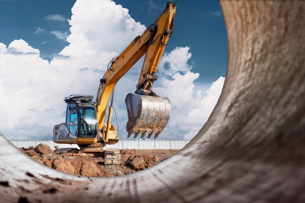 Un potente escavatore a cingoli scava il terreno contro il cielo blu lavori di sterro con attrezzature pesanti in cantiere vista da un grande tubo di cemento