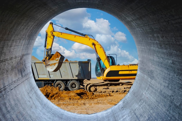 A powerful crawler excavator loads the earth into a dump truck against the blue sky Development and removal of soil from the construction site View through a large concrete pipe