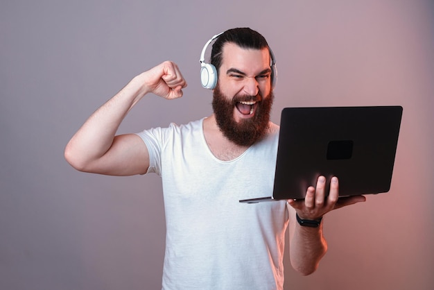 Powerful excited man wearing headphones is making the winner gesture while holding a laptop Studio shot over grey background with red light