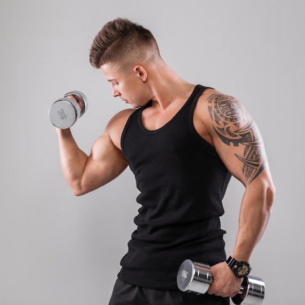 Powerful handsome fashionable young man with a muscular athletic body with tattoo in a black Tshirt workout with metal dumbbells on a gray background