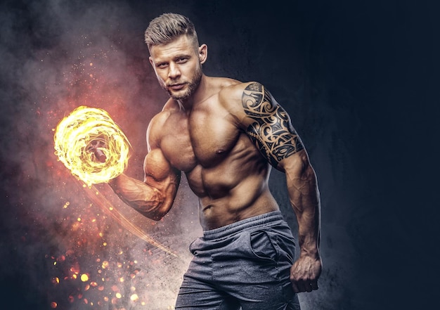 Powerful stylish bodybuilder with tattoo on his arm, doing the exercises on a biceps with dumbbells, look at the camera with a confident look. Fire art concept.