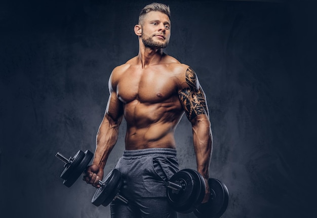 Powerful stylish bodybuilder with tattoo on his arm, doing the exercises with dumbbells. Isolated on a dark background.