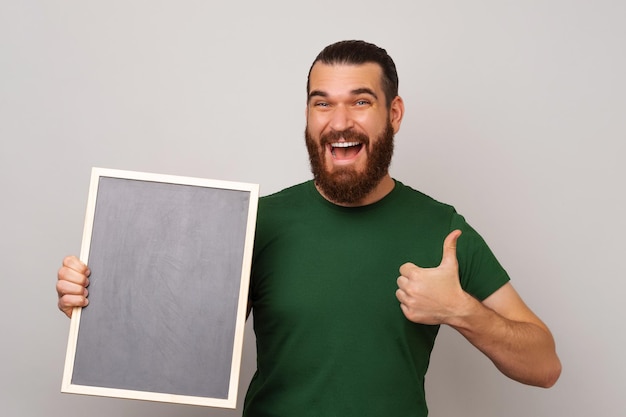 Powerful young bearded man is holding a small blackboard and thumb up