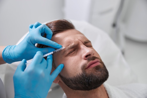注意深い美容師が眉間にリフティングフィラーを注射している間、患者の痛みのしかめっ面