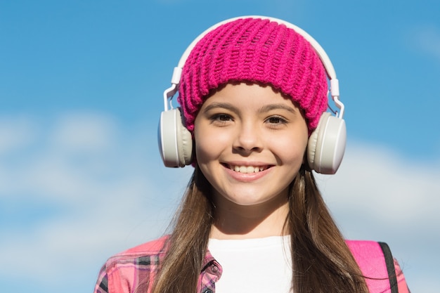 あなたの子供の耳を保護します。幸せな女の子は晴れた青い空にイヤホンを着用します。小さな子供はヘッドフォンで音楽を聴きます。耳と聴覚のケア。現代の生活。新技術。あなたを楽しませ、安全に保ちます。