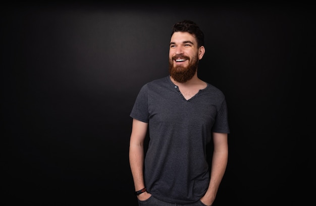 Pprtrait of bearded man standing over dark background  with hands in pockets while looking away