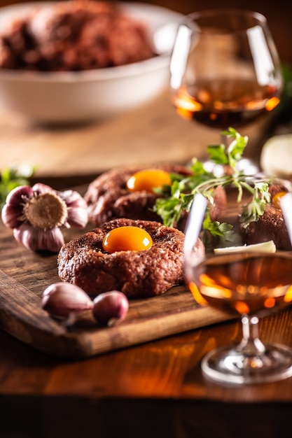 Prachtig bereide rundersteaks tartaar met smakelijke eidooiers. Geserveerd met knoflook, ui, kruiden en alcohol als drank. Witte kom vol rauw vlees op de achtergrond.