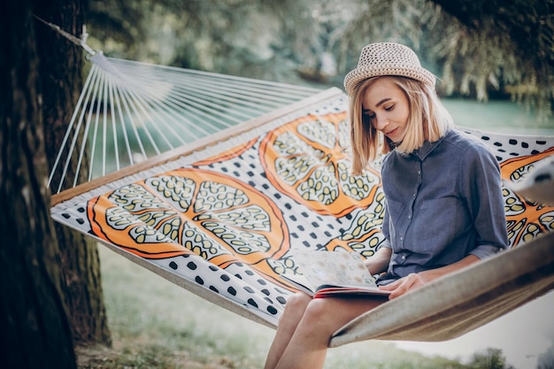 Foto prachtig blond hipster meisje in blauw shirt en vintage hoed die een boek leest op een hangmat jonge vrouw op een kampeertrip in het bosmeer op de achtergrond