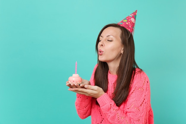 Prachtig jong meisje in gebreide roze trui verjaardag hoed met gesloten ogen uitblazen kaars op taart in handen geïsoleerd op blauwe achtergrond studio portret. Mensen levensstijl concept. Bespotten kopie ruimte.