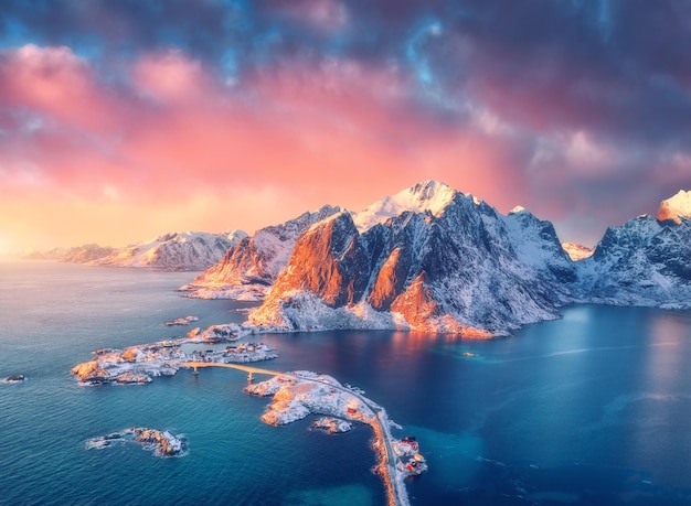 Prachtig landschap met blauwe zee besneeuwde bergen rotsen en eilanden dorp rorbu verkeersbrug en roze lucht bij zonsopgang Luchtfoto Hamnoy in de sneeuw in de winter op de Lofoten-eilanden Noorwegen Bovenaanzicht