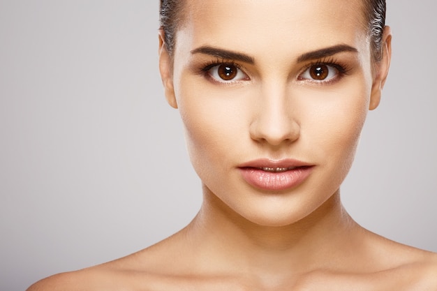 Prachtig meisje met bruin haar, schone huid en naakte schouders poseren bij grijze studio achtergrond, een model met lichte naakt make-up, close-up, portret.