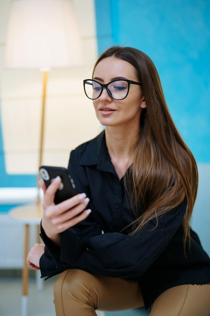 Prachtig mooi meisje lacht en poseert voor de camera in een moderne bril Vrouw zit en kijkt naar de smartphone Lichte interieurkleuren Close-up