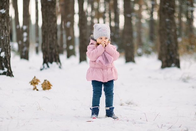 Prachtig winterportret in de buitenlucht van een schattig 4-jarig kindmeisje, met een stijlvolle grijze pet en roze jas, verwarmt haar handen en blaast erop. Frosty winterdag in besneeuwde bos.