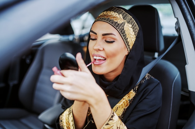 Prachtige aantrekkelijke jonge moslimvrouw in traditionele slijtage zittend in haar auto tijdens verkeersopstopping en lippenstift op te zetten.